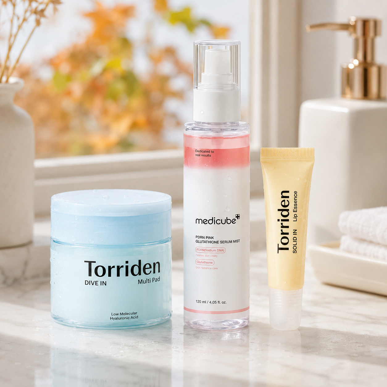 Three Torriden skincare products arranged on a marble surface in a bright bathroom setting: a light blue jar labeled Dive In Multi-Ph, a clear bottle with pink liquid labeled medicube Serum 9000, and a pale yellow tube labeled Torriden Advanced Retinol Treatment.